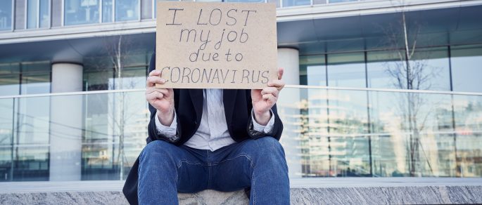 Man sitting outside of office building holding handmade sign stating that he has lost his job due to COVID-19